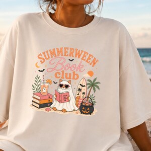 May include: An off-white short-sleeve t-shirt featuring a graphic print with the text "SUMMERWEEN Book club". The design includes a cat wearing a ghost costume and heart-shaped sunglasses, reading a book, surrounded by books, a drink, a surfboard, a palm tree, and a jack-o'-lantern bucket.