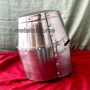 May include: A silver medieval helmet with a pointed top and a visor with a small opening for the eyes. The helmet has rivets and a textured surface.