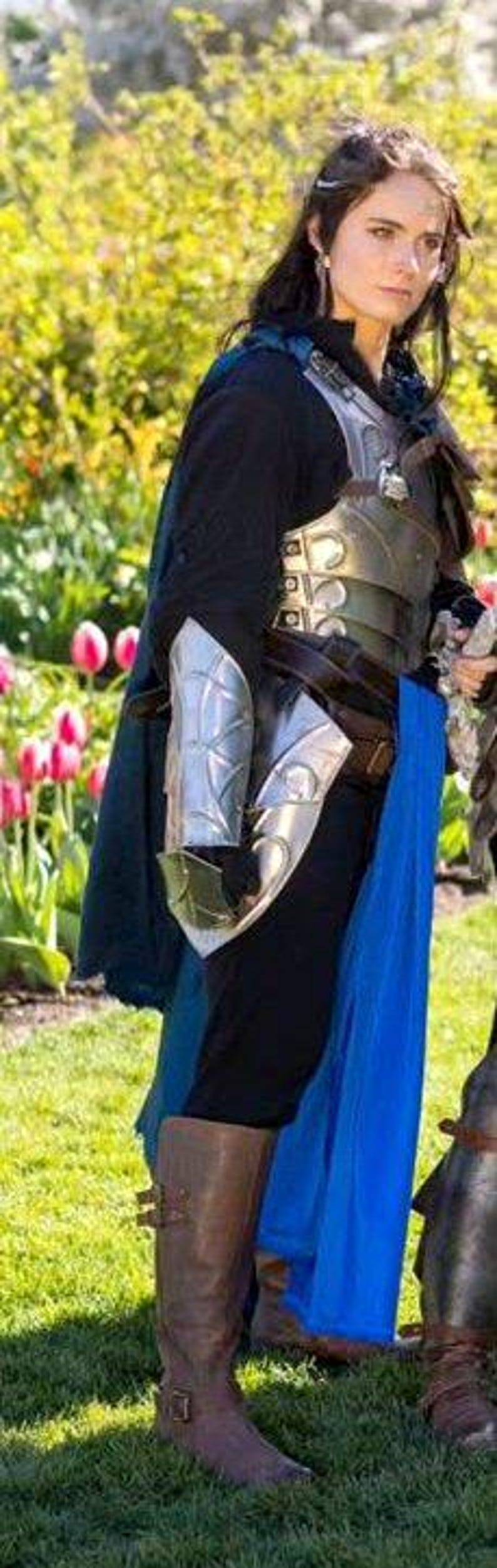 K&ouml;nnte beinhalten: Eine Frau in einem blau-schwarzen Kost&uuml;m mit silberner R&uuml;stung, einem blauen Band und braunen Stiefeln. Sie steht in einem Garten mit gr&uuml;nem Gras und rosa Blumen.