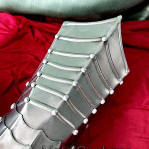 May include: Close-up of a silver metal gauntlet with a pointed design and rivets. The gauntlet is on a red fabric background.