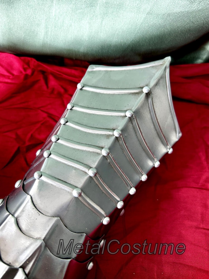May include: Close-up of a silver metal gauntlet with a pointed design and rivets. The gauntlet is on a red fabric background.