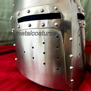 May include: A silver medieval helmet with a visor and a metal face mask. The helmet has a series of rivets around the edges and a small ventilation hole on the top. The helmet is made of metal and has a shiny finish. The text "metalcostume" is visible on the helmet.