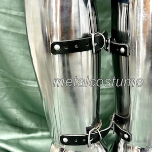 May include: A pair of silver metal shin guards with black leather straps and buckles. The shin guards are worn over jeans.