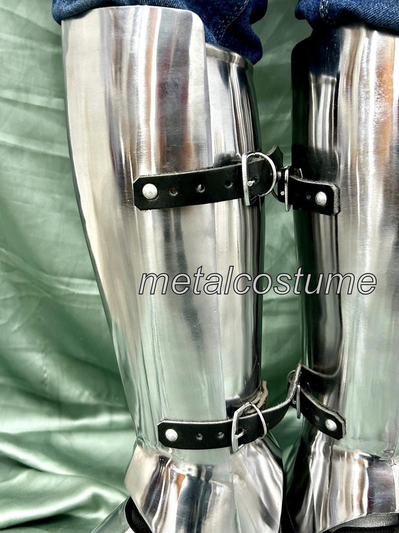 May include: A pair of silver metal shin guards with black leather straps and buckles. The shin guards are worn over jeans.