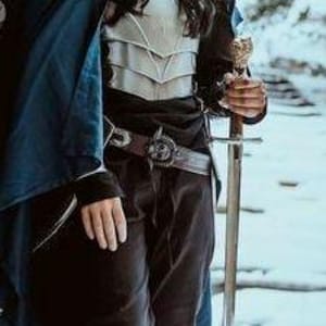 K&ouml;nnte beinhalten: Eine Frau in einem blauen Umhang und silberner R&uuml;stung h&auml;lt ein Schwert. Sie steht in einem verschneiten Wald.