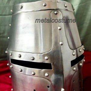 May include: A silver knight helmet with a visor and rivets. The helmet is made of metal and has a textured surface. The helmet is a replica of a medieval helmet.