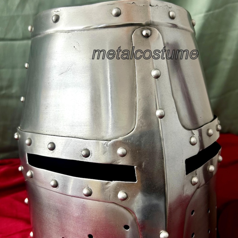 May include: A silver knight helmet with a visor and rivets. The helmet is made of metal and has a textured surface. The helmet is a replica of a medieval helmet.