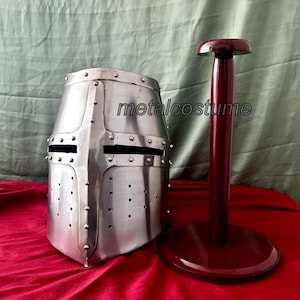 May include: A silver knight helmet with a visor and rivets, displayed on a dark red wooden stand. The helmet is made of metal and has a textured surface. The stand has a round base and a tall, cylindrical post.