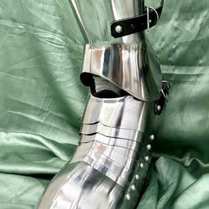 May include: Close-up of a silver metal shin guard with a black leather strap and buckle. The shin guard is made of multiple plates and has a pointed toe. The shin guard is on a green fabric background.