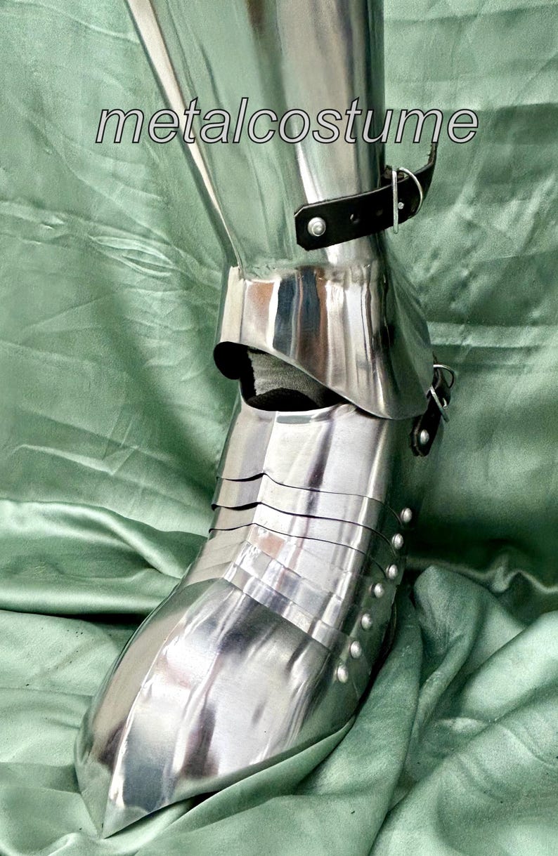 May include: Close-up of a silver metal shin guard with a black leather strap and buckle. The shin guard is made of multiple plates and has a pointed toe. The shin guard is on a green fabric background.