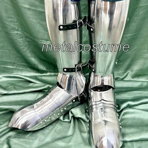 May include: A pair of silver metal shin guards with black leather straps. The shin guards are worn over jeans and have a pointed toe.