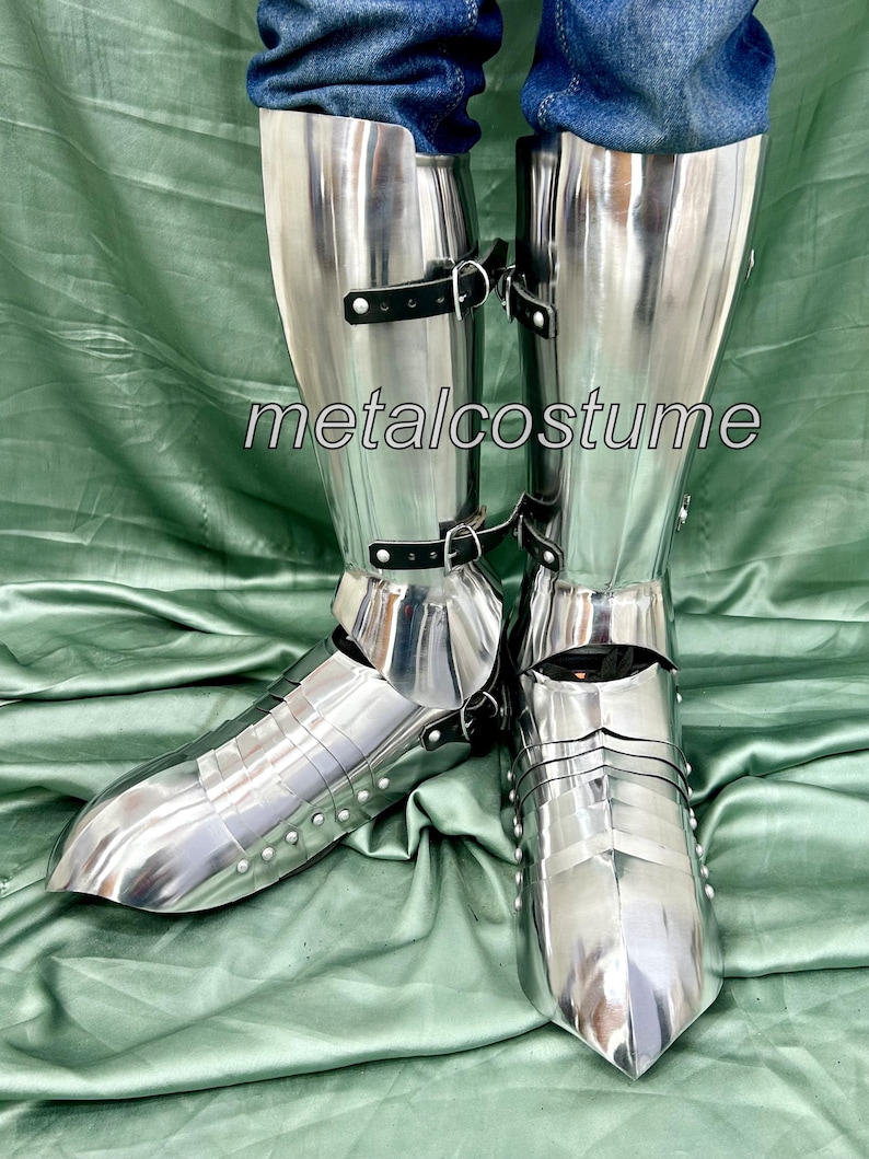 May include: A pair of silver metal shin guards with black leather straps. The shin guards are worn over jeans and have a pointed toe.