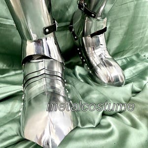 May include: A pair of silver metal knight's shin guards with buckles and straps. The shin guards are on a green fabric surface. The text "metalcostume" is visible in the image.