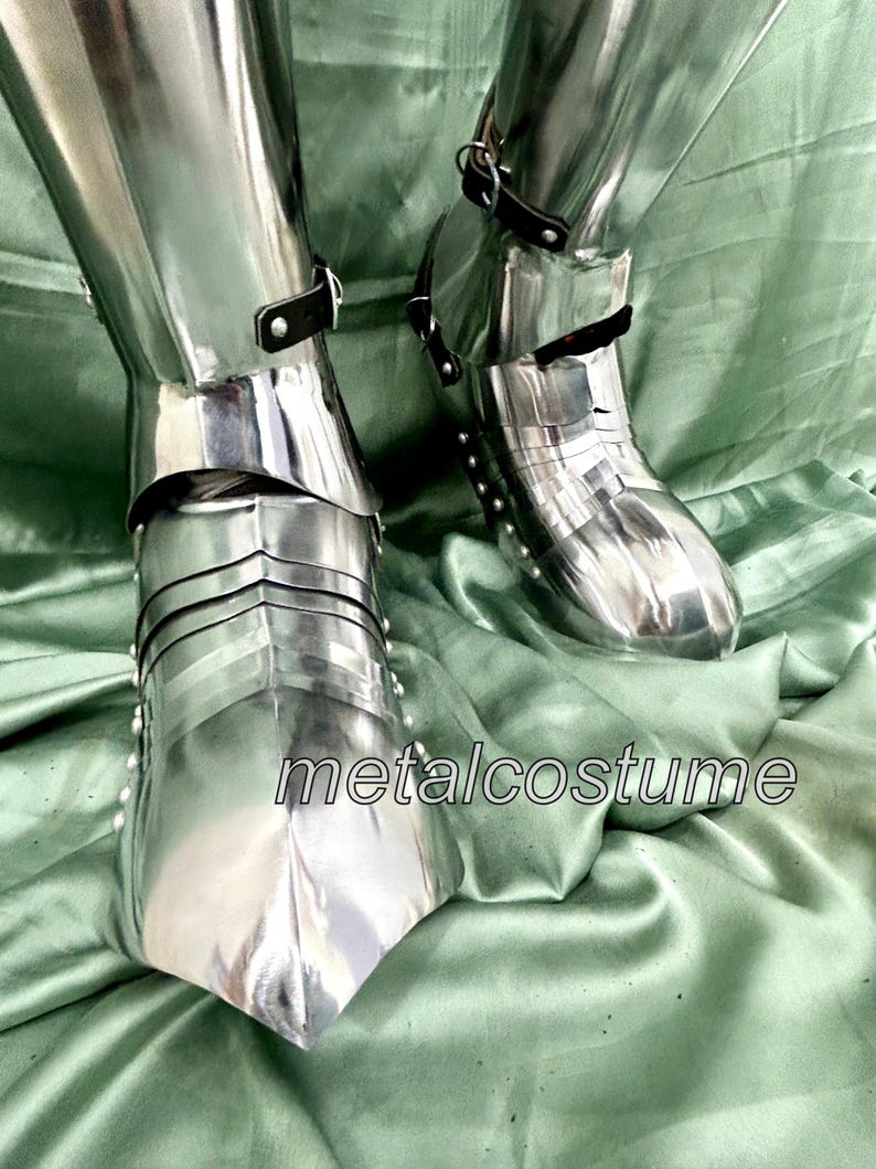 May include: A pair of silver metal knight's shin guards with buckles and straps. The shin guards are on a green fabric surface. The text "metalcostume" is visible in the image.