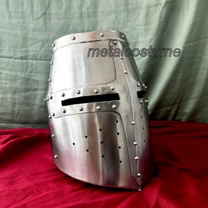 May include: A silver medieval knight helmet with a visor and rivets. The helmet is made of metal and has a textured surface. The helmet is on a red surface.