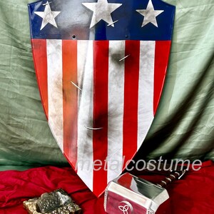 Puede incluir: Un escudo de Capit&aacute;n Am&eacute;rica rojo, blanco y azul con tres estrellas blancas sobre un fondo azul. El escudo est&aacute; hecho de metal y tiene un aspecto desgastado. Un martillo de Thor plateado con un mango de madera est&aacute; en primer plano. El martillo tiene un s&iacute;mbolo triquetra en la cabeza. Los objetos est&aacute;n sobre una superficie roja.