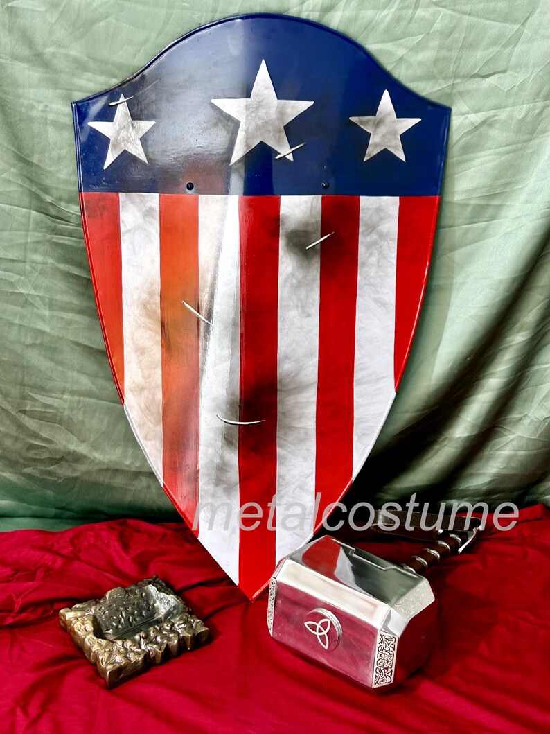 Puede incluir: Un escudo de Capit&aacute;n Am&eacute;rica rojo, blanco y azul con tres estrellas blancas sobre un fondo azul. El escudo est&aacute; hecho de metal y tiene un aspecto desgastado. Un martillo de Thor plateado con un mango de madera est&aacute; en primer plano. El martillo tiene un s&iacute;mbolo triquetra en la cabeza. Los objetos est&aacute;n sobre una superficie roja.