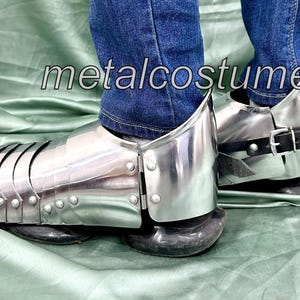 May include: A pair of silver metal shin guards with black leather straps. The shin guards are worn over jeans and have a medieval design.