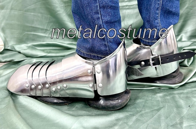 May include: A pair of silver metal shin guards with black leather straps. The shin guards are worn over jeans and have a medieval design.