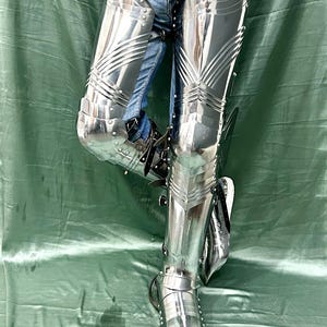 Op de afbeelding: Gepolijste stalen beenpantser met scharnierende platen en klinknagels. Het pantser is zeer reflecterend, met een puntig teenontwerp. De tekst "MetalCostume" is zichtbaar onderaan de afbeelding.