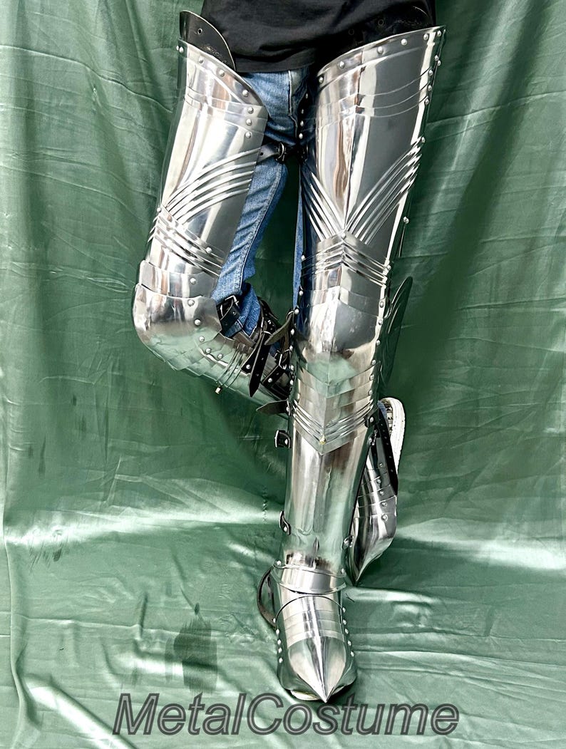 Op de afbeelding: Gepolijste stalen beenpantser met scharnierende platen en klinknagels. Het pantser is zeer reflecterend, met een puntig teenontwerp. De tekst "MetalCostume" is zichtbaar onderaan de afbeelding.