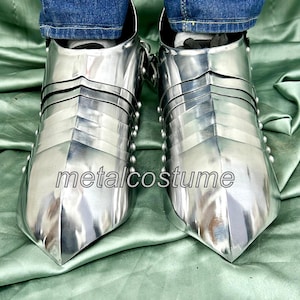 May include: A pair of silver metal medieval-style footwear with pointed toes. The footwear is made of metal plates and has a riveted design. The footwear is worn by a person wearing blue jeans.