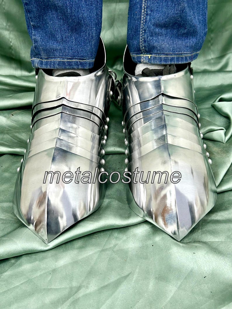 May include: A pair of silver metal medieval-style footwear with pointed toes. The footwear is made of metal plates and has a riveted design. The footwear is worn by a person wearing blue jeans.