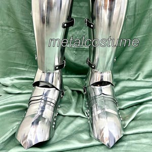 May include: A pair of silver metal knight's leg armor with black straps. The armor is made of metal plates and has a pointed toe.