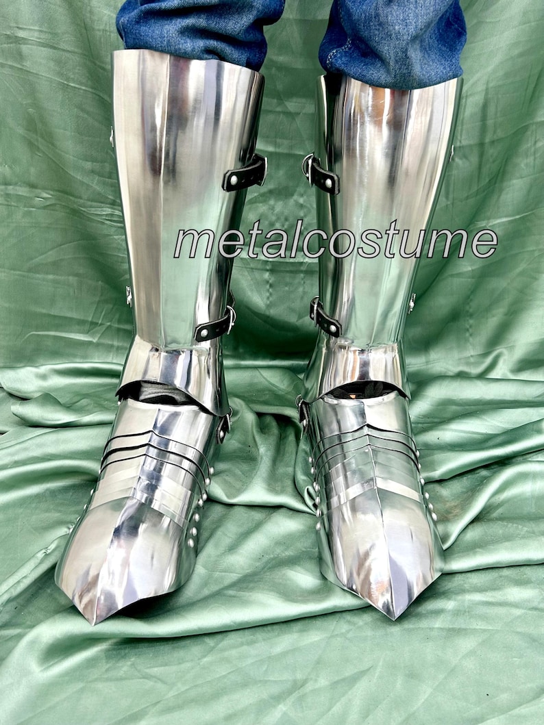 May include: A pair of silver metal knight's leg armor with black straps. The armor is made of metal plates and has a pointed toe.