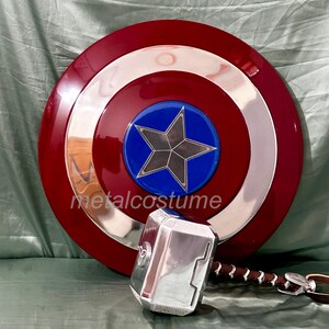 Op de afbeelding: Een rood, wit en blauw Captain America schild met een zilveren metalen rand en een blauwe ster in het midden. Een zilveren metalen hamer met een bruin leren handvat bevindt zich op de voorgrond.