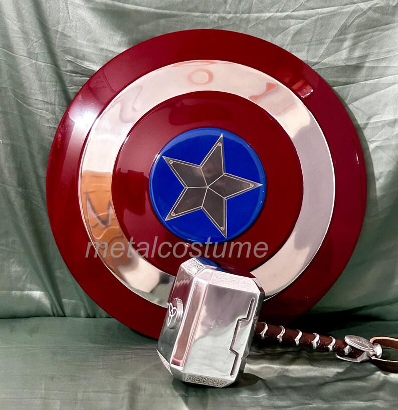 Op de afbeelding: Een rood, wit en blauw Captain America schild met een zilveren metalen rand en een blauwe ster in het midden. Een zilveren metalen hamer met een bruin leren handvat bevindt zich op de voorgrond.