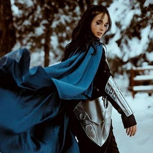K&ouml;nnte beinhalten: Eine Frau in einem blauen Umhang und silberner R&uuml;stung steht in einem verschneiten Wald. Der Umhang weht im Wind und die Frau schaut nach unten.