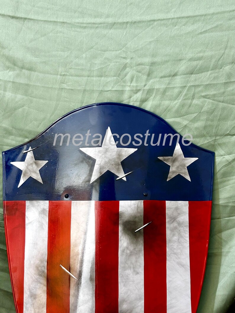 Puede incluir: Un escudo rojo, blanco y azul con tres estrellas blancas. El escudo est&aacute; hecho de metal y tiene un aspecto desgastado. El texto "metalcostume" es visible en el escudo.