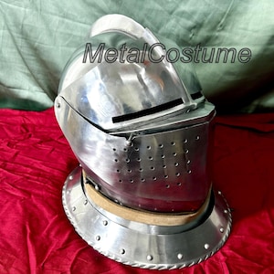 May include: A silver helmet with a visor and a leather strap. The helmet has a textured surface and a small metal crest on top. The helmet is on a red fabric background. MetalCostume is printed on the helmet.