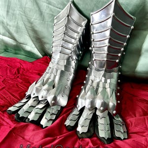 May include: A pair of silver metal gauntlets with a claw-like design. The gauntlets are made of interlocking metal plates and have a textured surface. The gauntlets are on a red fabric surface.