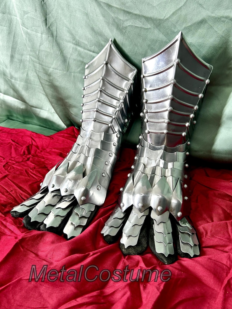 May include: A pair of silver metal gauntlets with a claw-like design. The gauntlets are made of interlocking metal plates and have a textured surface. The gauntlets are on a red fabric surface.