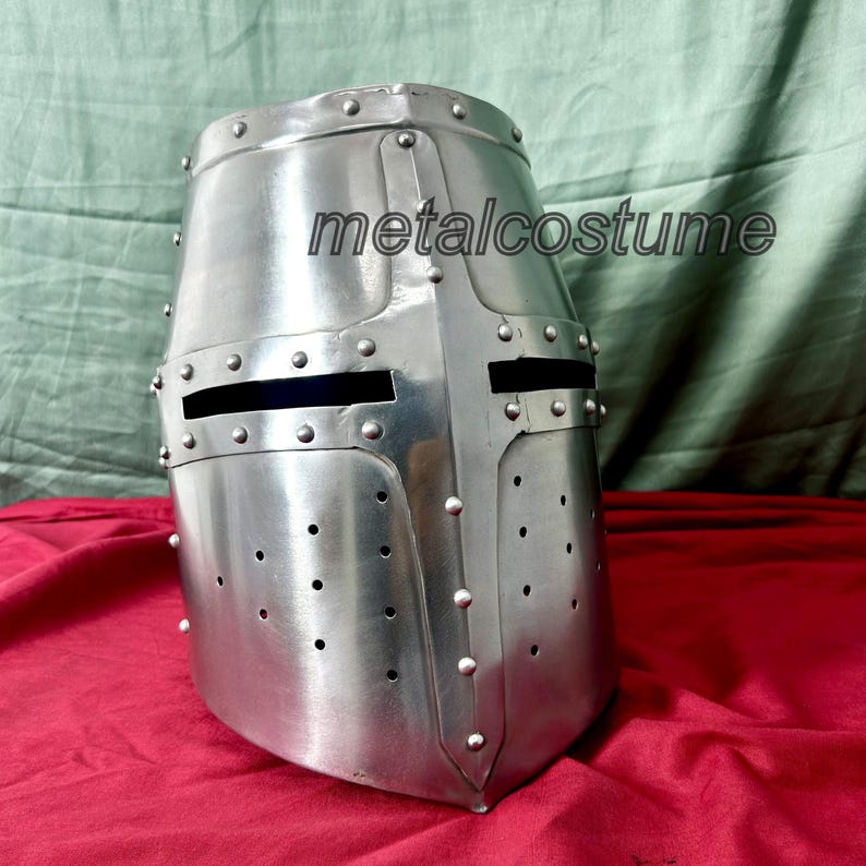 May include: A silver knight helmet with a visor and a metalcocostume logo. The helmet has a pointed top and a rounded bottom. It is decorated with rivets and has a small opening for the eyes.