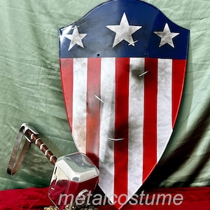 Puede incluir: Un martillo plateado con una correa de cuero marr&oacute;n y un escudo rojo, blanco y azul con estrellas. El escudo est&aacute; ligeramente desgastado y tiene un aspecto desgastado. El martillo y el escudo est&aacute;n sobre una superficie roja.