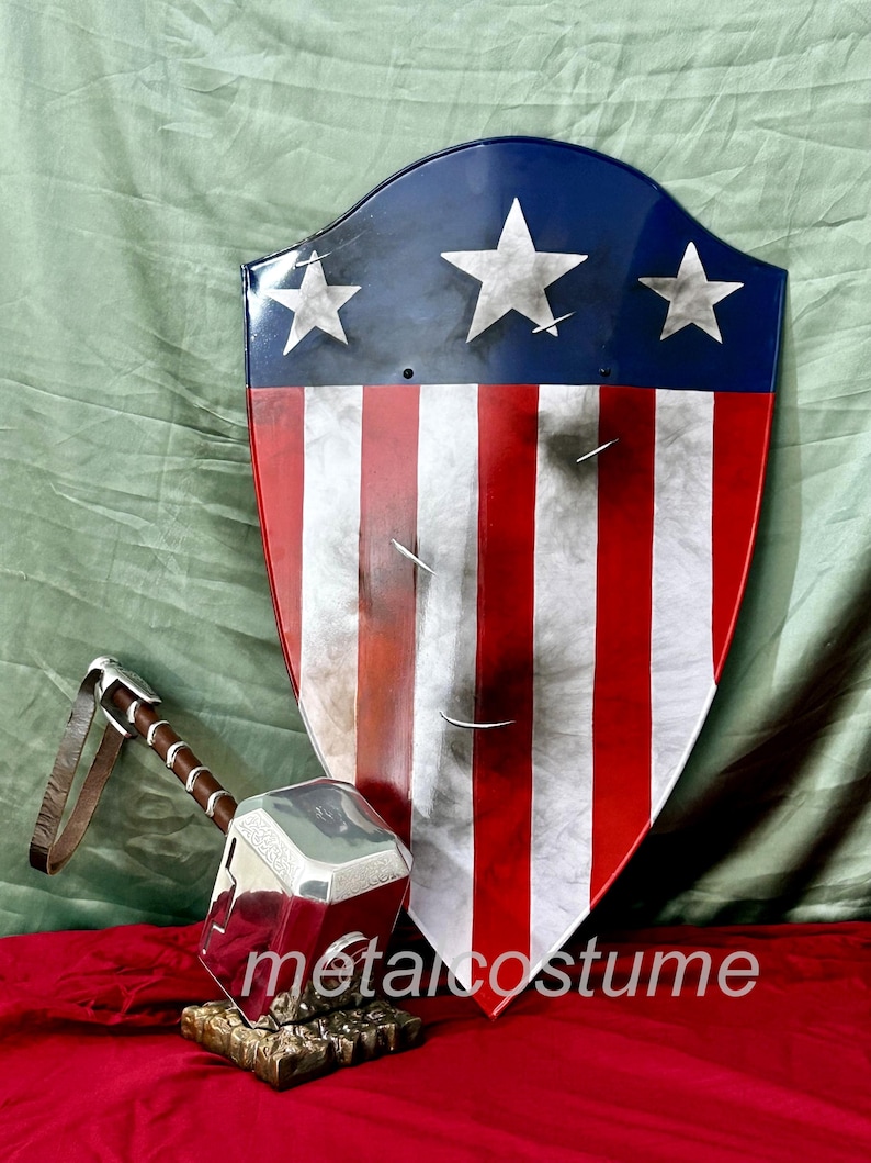 Puede incluir: Un martillo plateado con una correa de cuero marr&oacute;n y un escudo rojo, blanco y azul con estrellas. El escudo est&aacute; ligeramente desgastado y tiene un aspecto desgastado. El martillo y el escudo est&aacute;n sobre una superficie roja.