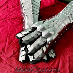 May include: A pair of silver metal gauntlets with a scaly design. The gauntlets are on a red fabric background. The gauntlets are closed and the fingers are curled inward. The gauntlets are made of metal plates that are riveted together. The gauntlets are a type of medieval armor.