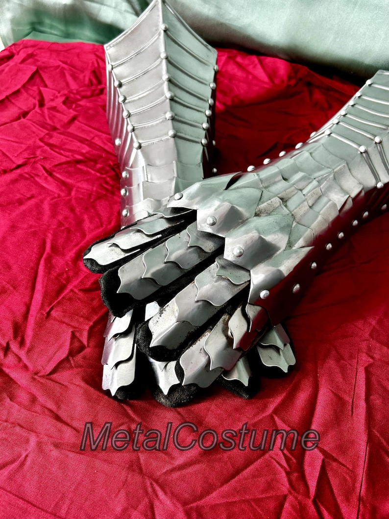 May include: A pair of silver metal gauntlets with a scaly design. The gauntlets are on a red fabric background. The gauntlets are closed and the fingers are curled inward. The gauntlets are made of metal plates that are riveted together. The gauntlets are a type of medieval armor.