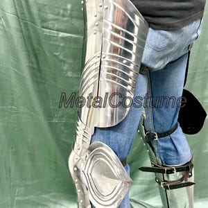 Op de afbeelding: Gepolijste zilveren metalen beenpantser met geklonken constructie en leren riemen. Het pantser is ontworpen om het been en de knie te beschermen. De tekst "MetalCostume" is zichtbaar op de afbeelding.