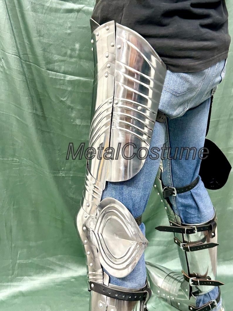 Op de afbeelding: Gepolijste zilveren metalen beenpantser met geklonken constructie en leren riemen. Het pantser is ontworpen om het been en de knie te beschermen. De tekst "MetalCostume" is zichtbaar op de afbeelding.
