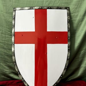 Könnte beinhalten: Ein weißer Schild mit einem roten Kreuz darauf bemalt. Der Schild ist aus Metall und hat einen dekorativen Rand. Der Schild ist im mittelalterlichen Stil gehalten und eignet sich perfekt für ein Kostüm oder eine Ausstellung.