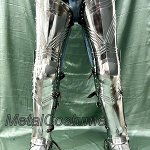 Op de afbeelding: Gepolijste metalen beenpantser met gelede platen en leren riemen. Het pantser is zeer reflecterend, met zichtbare klinknagels en een puntig teenontwerp. De tekst "MetalCostume" is zichtbaar. Dit pantser is waarschijnlijk voor historische re-enactment of kostuumgebruik.