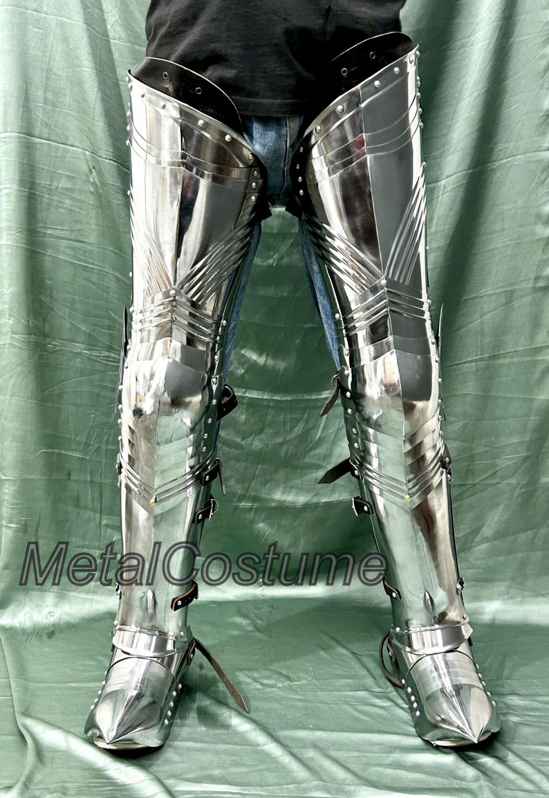 Op de afbeelding: Gepolijste metalen beenpantser met gelede platen en leren riemen. Het pantser is zeer reflecterend, met zichtbare klinknagels en een puntig teenontwerp. De tekst "MetalCostume" is zichtbaar. Dit pantser is waarschijnlijk voor historische re-enactment of kostuumgebruik.