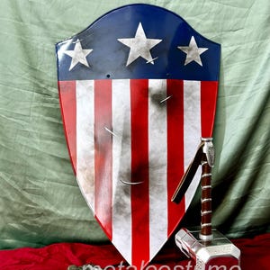Puede incluir: Un escudo rojo, blanco y azul con estrellas y rayas, y un martillo plateado con mango marr&oacute;n. El escudo est&aacute; ligeramente desgastado y tiene un aspecto envejecido. El martillo tiene detalles intrincados y una banda de metal alrededor del mango.