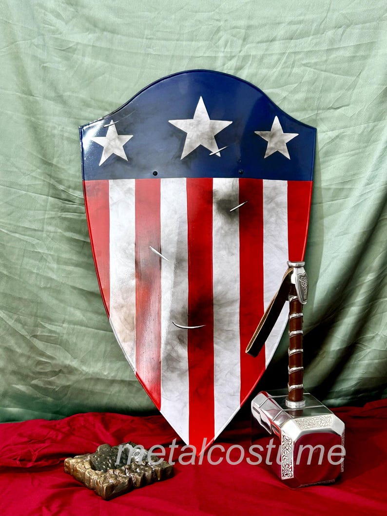 Puede incluir: Un escudo rojo, blanco y azul con estrellas y rayas, y un martillo plateado con mango marr&oacute;n. El escudo est&aacute; ligeramente desgastado y tiene un aspecto envejecido. El martillo tiene detalles intrincados y una banda de metal alrededor del mango.