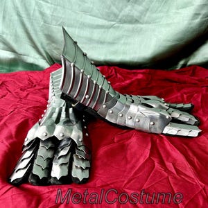 May include: A pair of silver metal gauntlets with a scaly design on the fingers and a ridged design on the forearm. The gauntlets are lying on a red fabric surface.