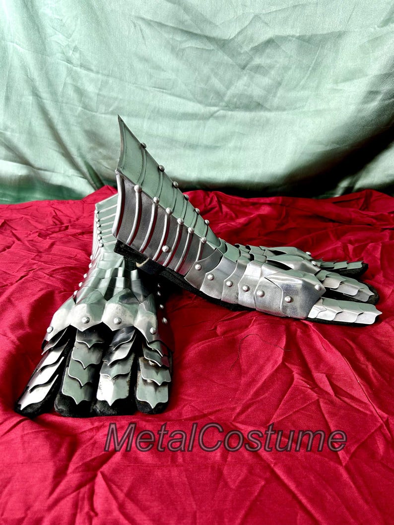 May include: A pair of silver metal gauntlets with a scaly design on the fingers and a ridged design on the forearm. The gauntlets are lying on a red fabric surface.